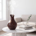 A dark brown wooden humidifier emitting a plume of steam, styled on a white marble round coffee table next to a stack of books and a small glass vase with a eucalyptus sprig, in front of a beige sofa.