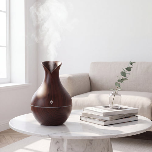 A dark brown wooden humidifier emitting a plume of steam, styled on a white marble round coffee table next to a stack of books and a small glass vase with a eucalyptus sprig, in front of a beige sofa.