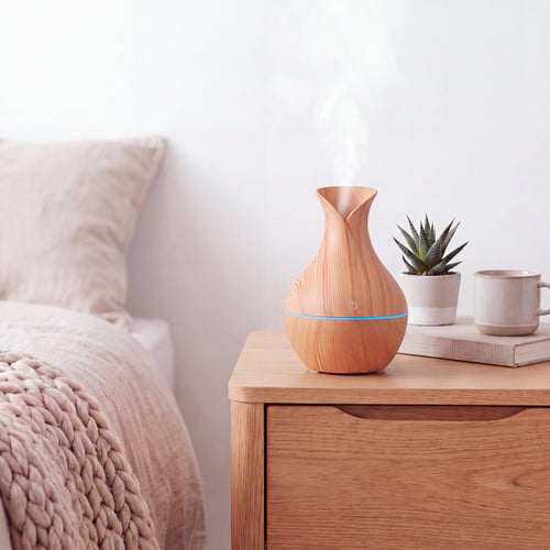 A wooden essential oil diffuser emitting a soft white mist, styled on a light wood bedside table next to a bed with a beige blanket, a small potted succulent, and a white mug.